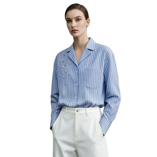 A model wearing an AMII light blue and white striped button-down shirt with pearl embellishments on the chest, paired with white trousers, set against a neutral gray background.