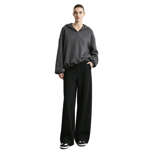 A model stands against a light gray wall wearing a dark gray half-zip hoodie, black wide-leg pants, and black-and-white sneakers.