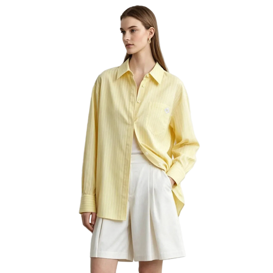 A woman modeling a pale yellow long-sleeve button-up shirt with subtle vertical stripes, worn open over a white top and white shorts. The AMII brand logo is visible in the top left corner against a light gray studio background.