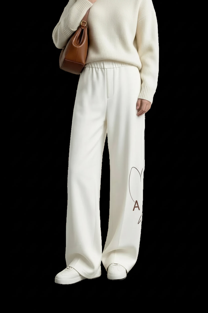 A model wearing AMII cream-colored wide-leg knit pants with a small heart and letter graphic on the lower left leg, styled with a cream sweater, white sneakers, and a brown shoulder bag against a minimalist studio background.