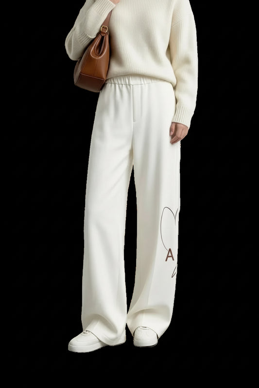 A model wearing AMII cream-colored wide-leg knit pants with a small heart and letter graphic on the lower left leg, styled with a cream sweater, white sneakers, and a brown shoulder bag against a minimalist studio background.