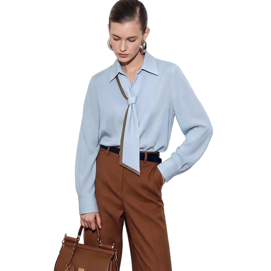 A woman models a light blue long-sleeve blouse with a tie-neck detail, paired with brown trousers and holding a brown handbag, set against a white paneled door. The brand name AMII is visible in the top left corner.